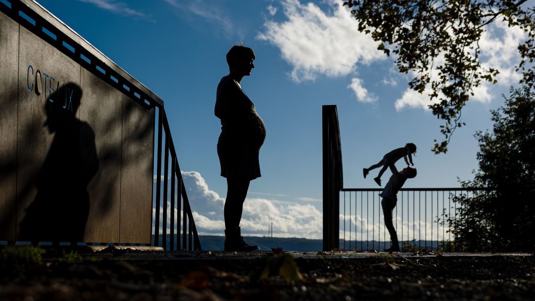 Photo de la doula r'Eve enceinte tenant son ventre et en arrière plan un homme portant un enfant dans les airs