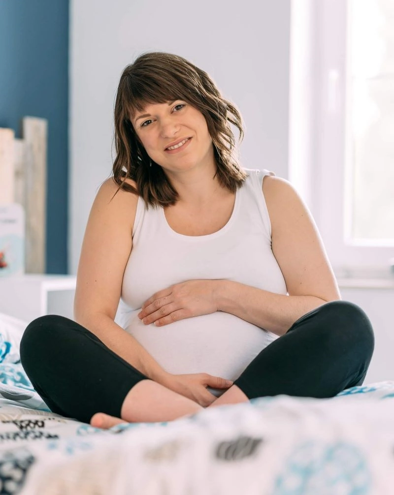 Photo de la doula r'Eve enceinte assise sur un lit regardant et tenant son ventre rond