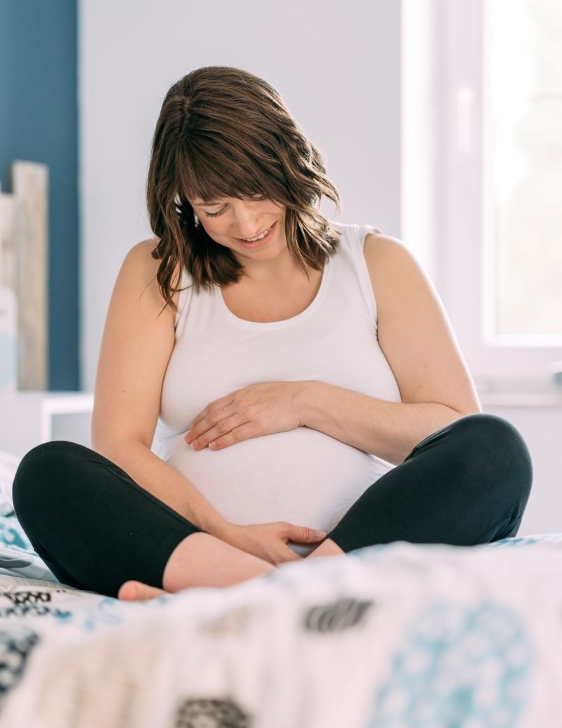 Photo de la doula r'Eve enceinte assise sur un lit regardant et tenant son ventre rond